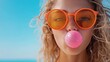 ©  Shomixer - A smiling young woman blowing bubble gum while wearing designer orange sunglasses, radiating confidence against a clear blue backdrop, highlighting a casual summer mood.