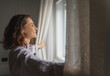© olezzo - Young relaxed woman standing at window open curtains in the morning