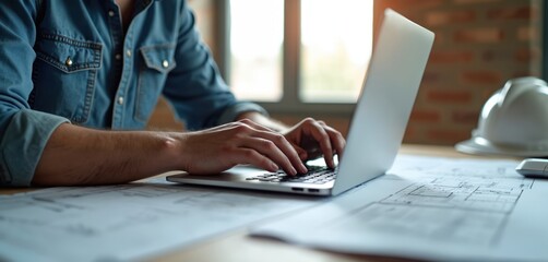 Man types on laptop keyboard at desk with blueprints. Engineering plans, architectural design, white hard hat visible. Engineer works on project in office, using tech to manage construction.