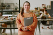 © AnnaStills - Portrait of smiling female office worker standing in office and holding gray clipboard
