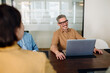 © Vadim Pastuh - Smiling businessman wearing glasses and a brown shirt, sitting at a conference table with a laptop. He engages in a professional meeting in a modern office setting.