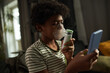 © pressmaster - African American youngster with oxygen mask on nose inhaling vapors and looking at smartphone screen in front of camera