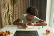 © pressmaster - Diligent African American pupil taking notes in copybook after teacher during online lesson while sitting in front of laptop