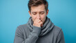 © Cathy - Young ill sick man wear gray sweater scarf cough sneeze cover mouth with hand isolated on plain blue background studio portrait. Healthy lifestyle disease virus treatment cold season recovery concept.