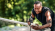 © Prostock-studio - Smiling african american sporty guy chatting on phone, drinking protein, exercising at park, empty space