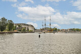 A picturesque river scene featuring historical ships alongside modern buildings, framed by lush greenery and a vibrant sky.