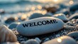 © meristock - Engraved welcome text on smooth light gray pebble positioned prominently on sandy beach, soft focus background with warm bokeh from golden sunset.