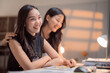 © MINAE - Two young asian businesswomen are working together in a modern office, one of them is putting a hand on the shoulder of her colleague, they are smiling and looking away
