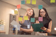 © MINAE - Two asian businesswomen sharing ideas using colorful sticky notes and writing on clipboard, working together on new project plan in modern office at night