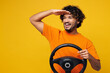 © ViDi Studio - Young smiling happy fun Indian man he wear orange t-shirt casual clothes hold steering wheel driving car look far away distance isolated on plain yellow background studio portrait. Lifestyle concept.