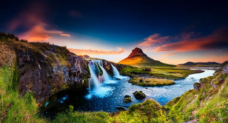 Poster - A breathtaking vista of a powerful waterfall cascading into a serene river, with a striking mountain silhouette against a vibrant sunset sky. Lush green grass and rugged rocks surround the scene.