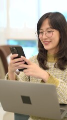 Fototapete - Vertical video of Asian woman smiling and wearing glasses while using smartphone to search internet, sitting at desk with laptop in bright indoor setting, enjoying communication and technology.