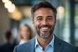 © angelmaxmixam - Close up of smiling man with beard wearing a suit in modern office setting. Two people are blurred in the background and light is soft.