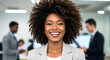 © Nedrofly - A afro confident executive stands at the forefront of a contemporary office, showcasing a vibrant smile. In the background, colleagues engage in discussions, emphasizing teamwork and leadership.