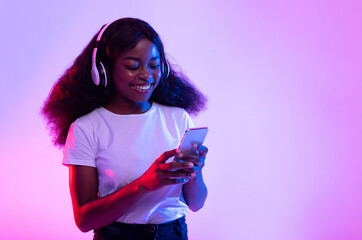 Poster - Cheerful millennial black female in wireless headphones using smartphone in neon light, copy space. Pretty young African American woman checking new mobile music app