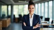 © saranya - A confident businesswoman stands with arms crossed, smiling in a modern office with colleagues working in the background