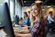 © MT - A Young Caucasian Woman Smiling at a Computer Desk in a Modern Office Space Surrounded by Colleagues Working on Their Computers