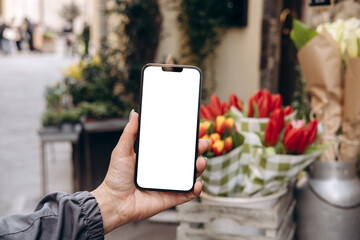 Mockup woman's hand holding using smart phone with white blank screen POV at flower street shop, mobile app design and advertising, online marketing