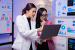 © Tj - Two asian businesswomen are discussing and analyzing financial charts and graphs displayed on a laptop, collaborating on a project in a modern office environment with a whiteboard and pinned charts
