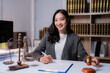 © Tj - Asian female lawyer smiling and signing legal document with gavel and Statue of Liberty figurine on the desk in law office, legal, justice and lawyer concept