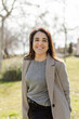 © BASILICOSTUDIO STOCK - A middle-aged Caucasian woman smiles while standing in a sunlit park. She looks confident and relaxed. Vertical image, natural light, blurred urban background with trees and greenery.
