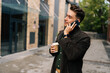 © dikushin - Handsome bearded young man drinking tasty coffee and talking on smartphone while walking in city street by modern urban buildings, enjoying pleasant conversation during break outdoors alone.