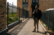 © dikushin - Full length portrait of handsome young man holding coffee cup standing by residential building with metal fence and brick walls, smiling looking away, enjoying peaceful moment outdoors.