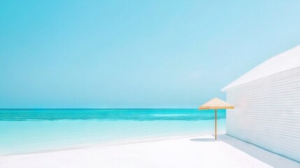Wall Mural - an umbrella sits on the beach adjacent to a white building and a blue ocean behind, with a blue sky 