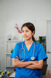 © Satori Studio - A focused female nurse in blue scrubs stands with arms crossed, wearing a stethoscope, in a contemporary medical office environment.