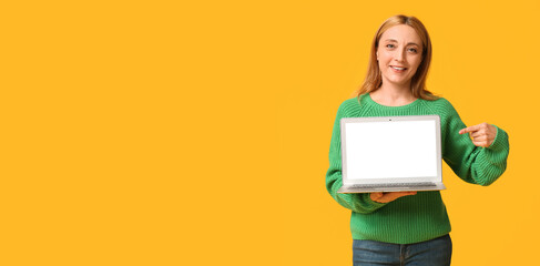Beautiful mature woman in stylish green sweater pointing at modern laptop on yellow background