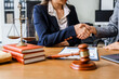 © Phushutter - Two female lawyers in business suits shake hands at wooden desk, finalizing legal agreement. wooden gavel and scales represent justice as they discuss corporate law, compliance, dispute resolution.