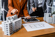 © Phushutter - Two businesswomen sit at a desk in a meeting, discussing property development while building a model of city tower. real estate investments, zoning laws, sustainable energy solutions. net zero