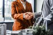 © Phushutter - Two businesswomen shake hands at desk, finalizing deal on property development. real estate investments, zoning laws, sustainable energy solutions while working on city tower model, net-zero goals.
