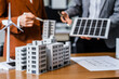 © Phushutter - Two businesswomen sit at a desk in a meeting, discussing property development while building a model of city tower. real estate investments, zoning laws, sustainable energy solutions. net zero