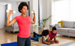 © NDABCREATIVITY - Young woman, mother exercising at home in living room, father playing with kid in background.