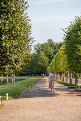Leinwandbilder - Woman walking along tree-lined path near pond in Peterhof during sunny day, enjoying the serene landscape and fresh air in late summer