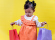 © Iqy - A stylish asian little girl in an orange dress holds red and purple shopping bags against a bright yellow background.