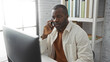 © Krakenimages.com - Young black man in office setting talking on phone while working on computer, showcasing a modern workplace environment.