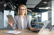 © Liubomir - Smiling woman in a blazer works at her desk with a laptop and tablet in a bright modern office with glass walls.