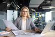 © Liubomir - Businesswoman working at her desk with a laptop, documents, and wearing glasses in a modern office environment.