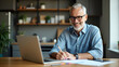 © Netaya - Happy smiling middle aged older mature man wearing glasses looking at laptop using computer writing notes watching webinar sitting at home table hybrid work elearning online learning concept. concept