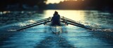 Rear view of a single rower in the early morning gleam, cutting through serene waters creating ripples in golden light.