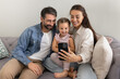 © fizkes - Happy mom and dad holding cute daughter kid on lap, resting on home sofa, using smartphone for online communication, video call talk, taking selfie, smiling, enjoying leisure with technology