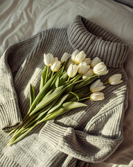 Poster - A still life photograph featuring a bouquet of white tulips placed atop a textured gray sweater, set against a backdrop of a white bedspread. The sweater's intricate weave and the delicate petals of t