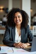 © JetHuynh - A smiling businesswoman with curly hair sits at a laptop in an office, colleagues blurred in the background. She's wearing a blazer and white shirt