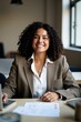 © JetHuynh - A smiling businesswoman with curly hair sits at a laptop in an office, colleagues blurred in the background. She's wearing a blazer and white shirt
