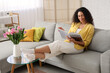 © Pixel-Shot - Young African-American woman with tulips reading magazine at home on spring day