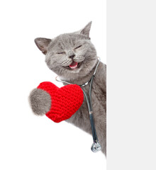 Fototapete - Happy cat dressed like a doctor with stethoscope on his neck looks from behind blank white banner and holds red heart. isolated on white background