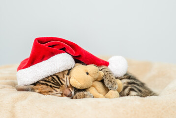 Aufkleber - Cute Bengal kitten wearing red santa's hat sleeps hugs favorite toy bear. Empty space for text