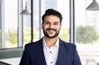 © fizkes - Young Indian male project leader dressed in formal suit posing in modern coworking, looking at camera with smile, exudes success and professionalism, office portrait. Career, achievements, leadership